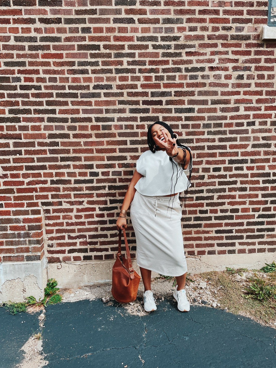 Black woman in a cream shirt and skirt throwing up the peace sign in front of brick wall