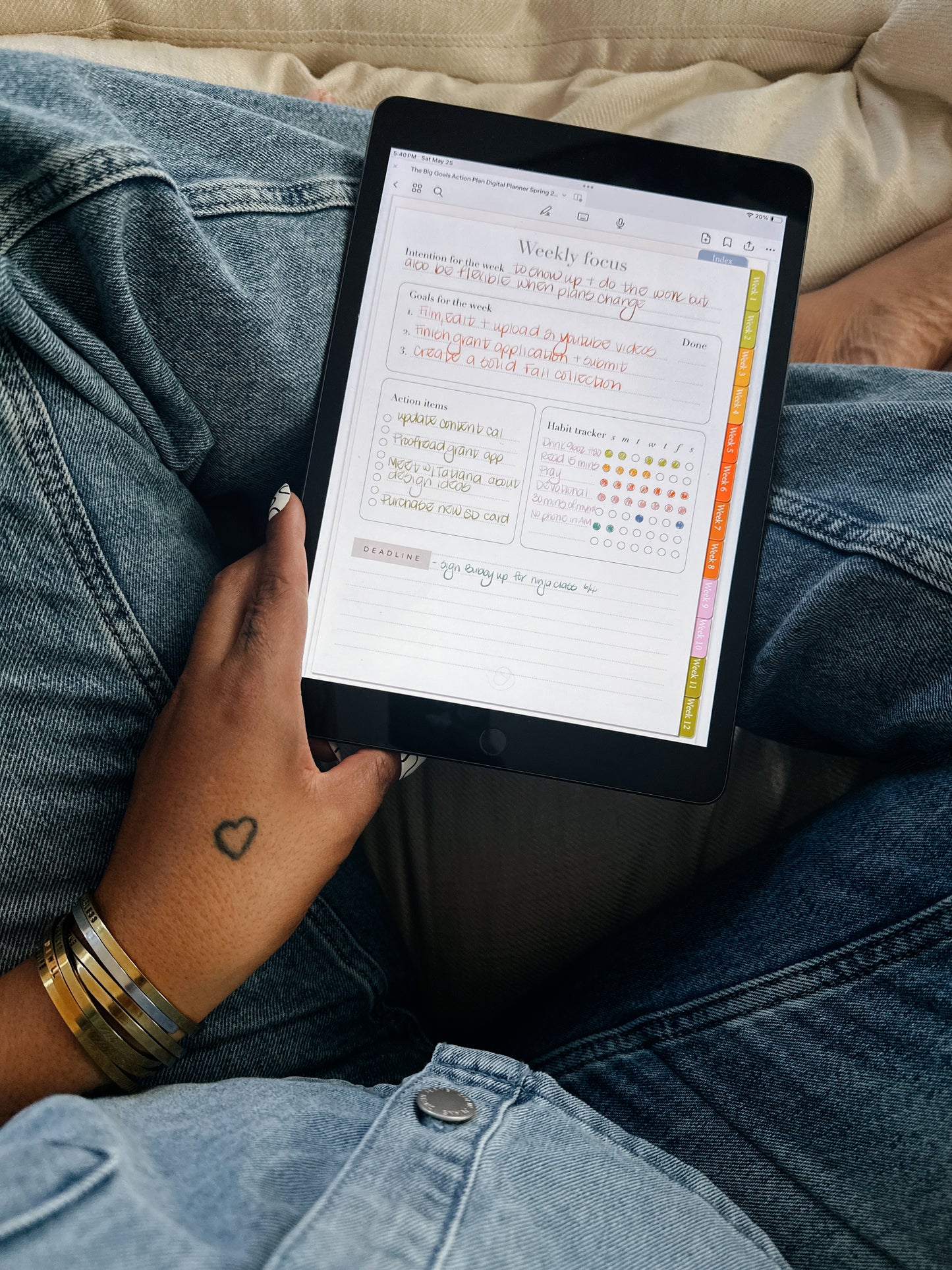 A person holding an iPad displaying the 'Weekly Focus' section of 'The Big Goals Action Plan' planner while sitting cross-legged in jeans.