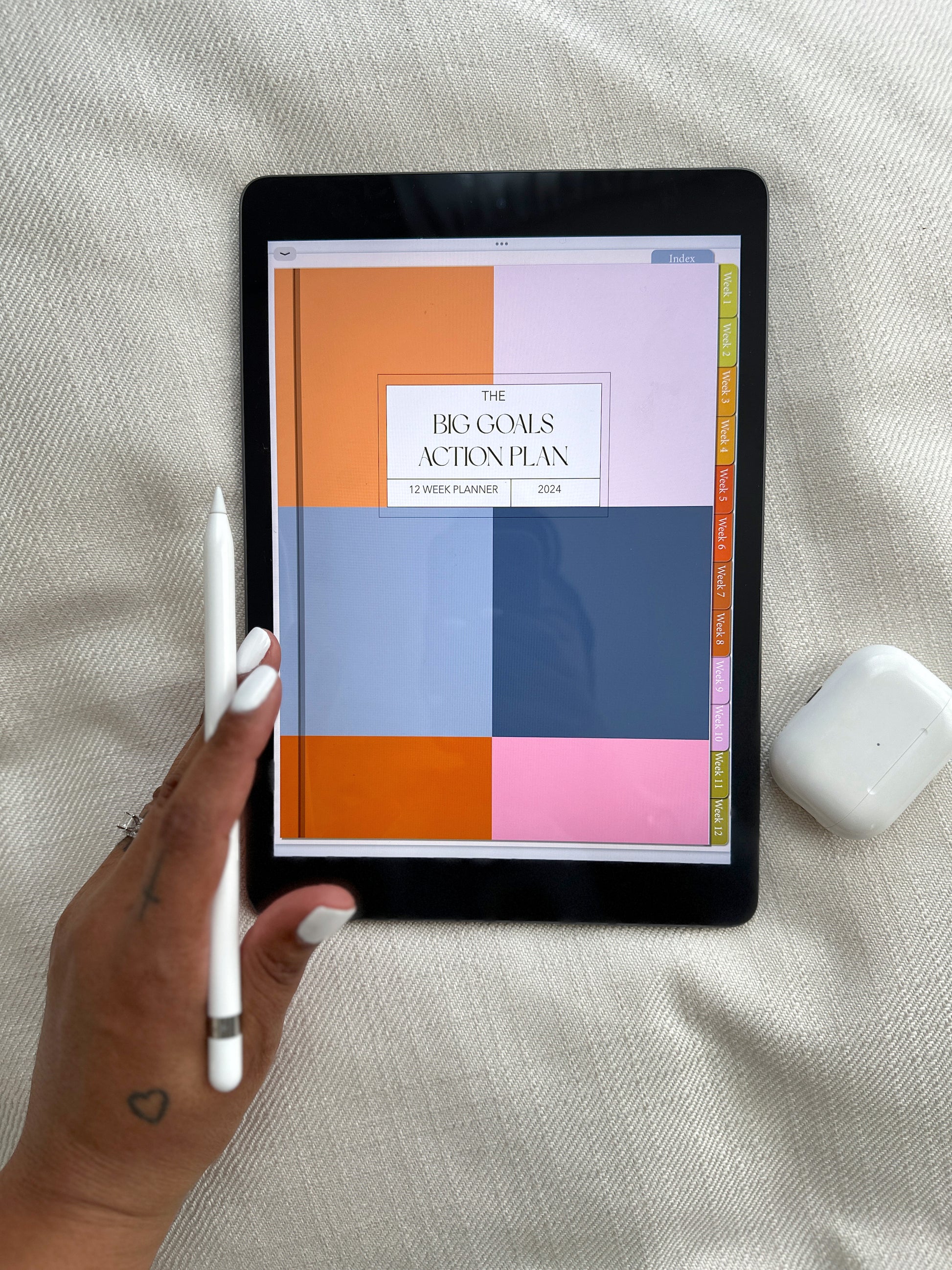 A person using an Apple Pencil on an iPad displaying 'The Big Goals Action Plan' 12-week planner for 2024. The colorful planner has week labels on the right. AirPods are visible beside the iPad.