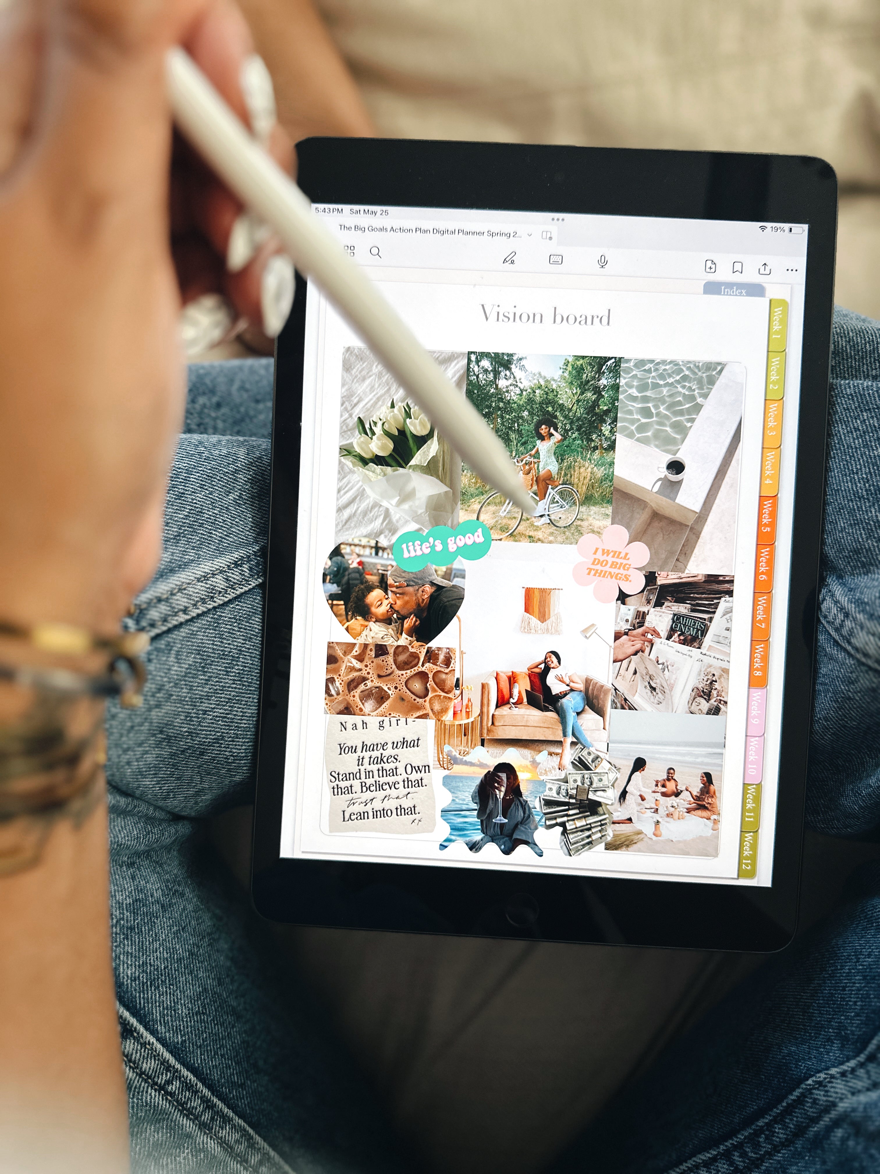 A person using an Apple Pencil to interact with an iPad displaying a 'Vision Board' section of 'The Big Goals Action Plan' planner. The vision board includes various motivational images and quotes. The iPad is held in the person's lap, who is wearing jeans.